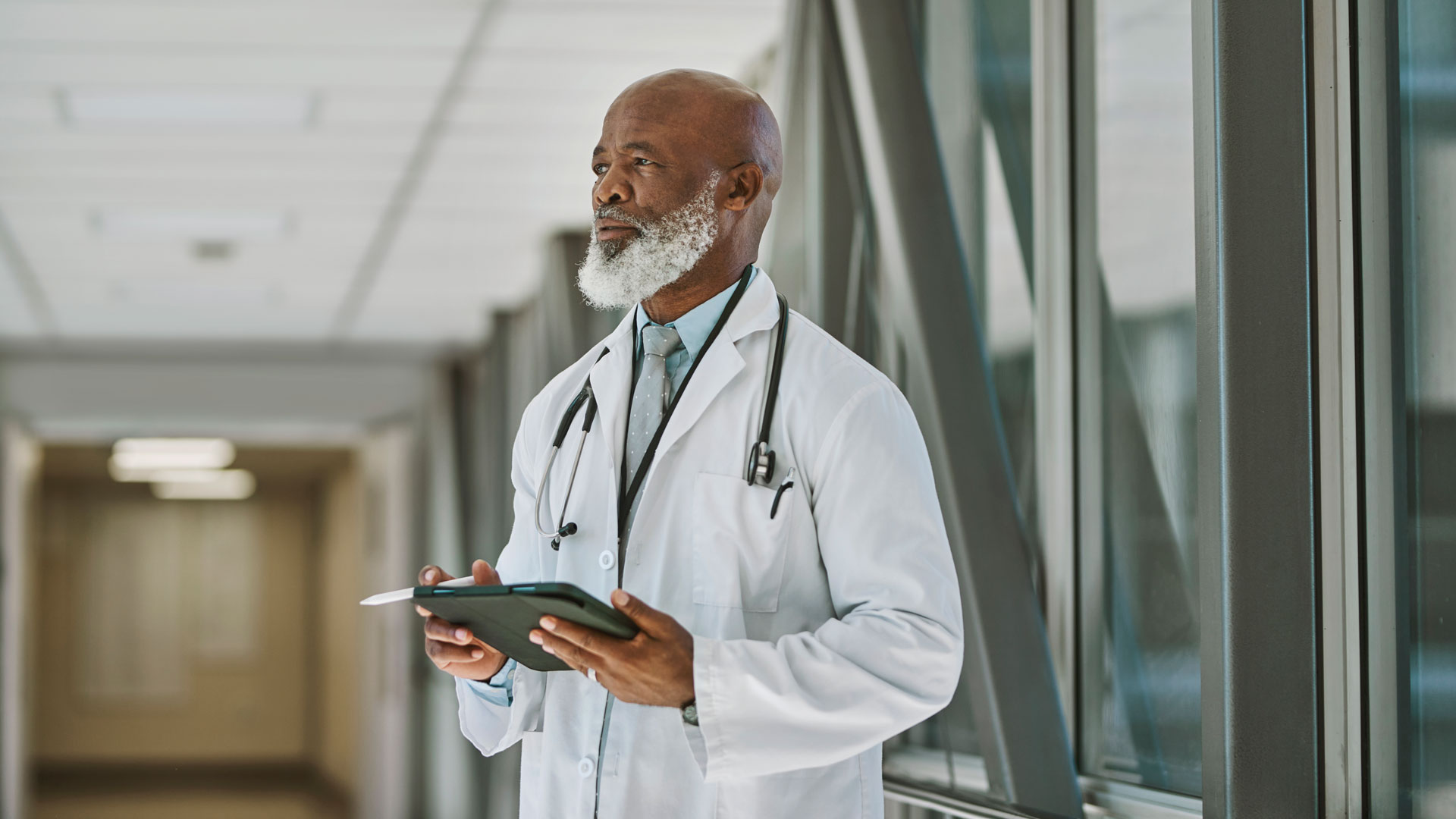 Senior doctor with white beard in contemplation, holding digital tablet and looking into distance at hospital corridor.